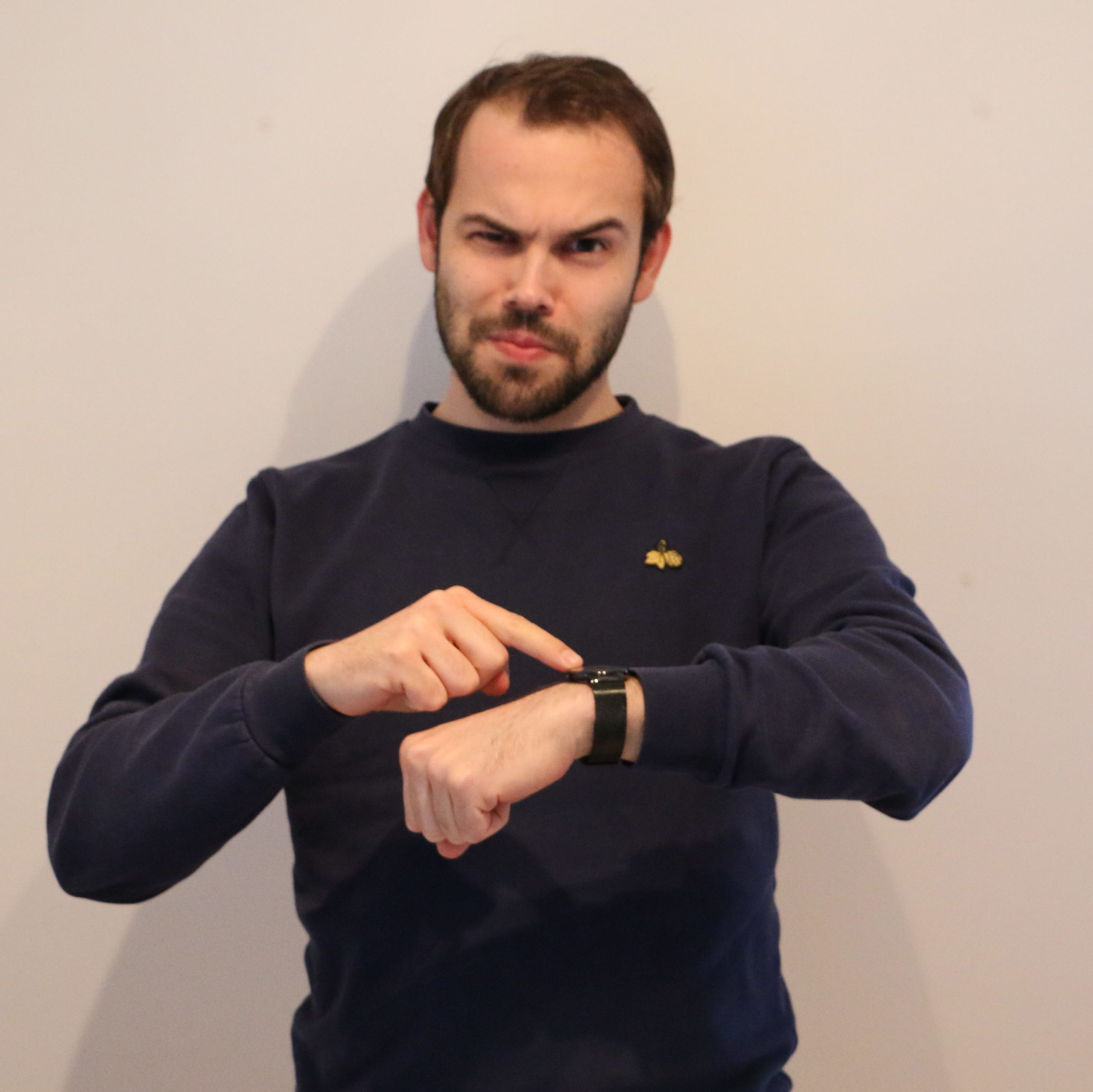 Benedikt Migge blickt grimmig-belustigt in die Kamera. Mit der rechten Hand zeigt er auf seine Armbanduhr.